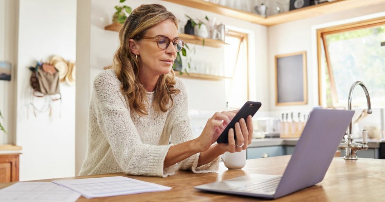 Mature woman sitting at kitchen counter using smartphone and laptop